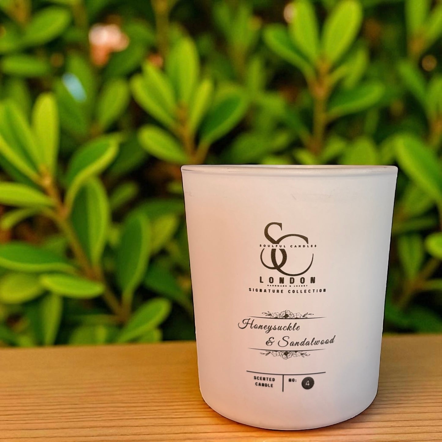 White candle with branding and scent description on a wooden surface with green foliage in the background