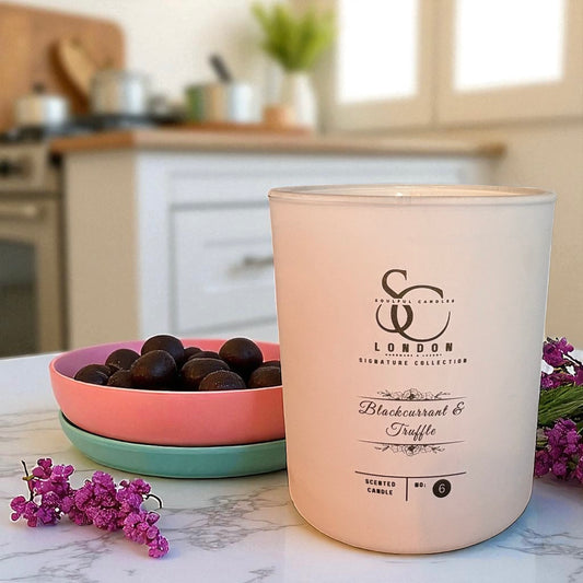 Candle with branding on a kitchen counter with a bowl of blackberries and purple flowers.