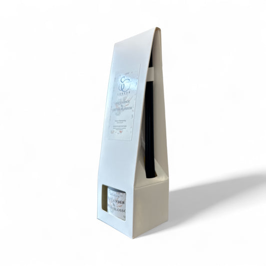 Angled view of Soft Amber & Cotton Blossom 100ml odour eliminating reed diffuser in white box, showing label and black reeds, handmade in London