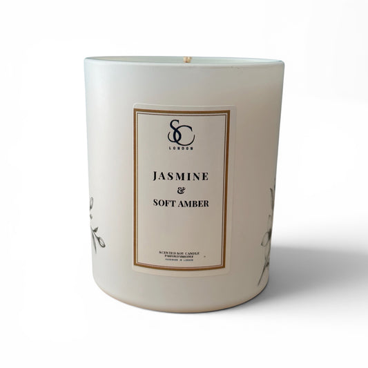 A large white soy candle in a cylindrical glass jar with a cream-colored front label. The label features a thin gold border and black text reading 'SC London JASMINE & SOFT AMBER'.