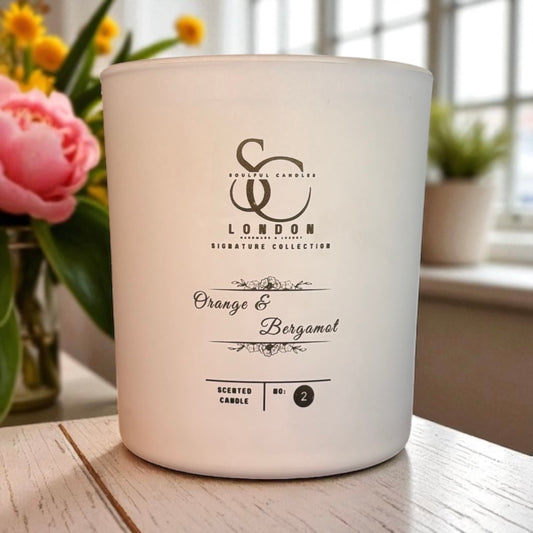 White candle with branding and fragrance description on a wooden surface with flowers in the background
