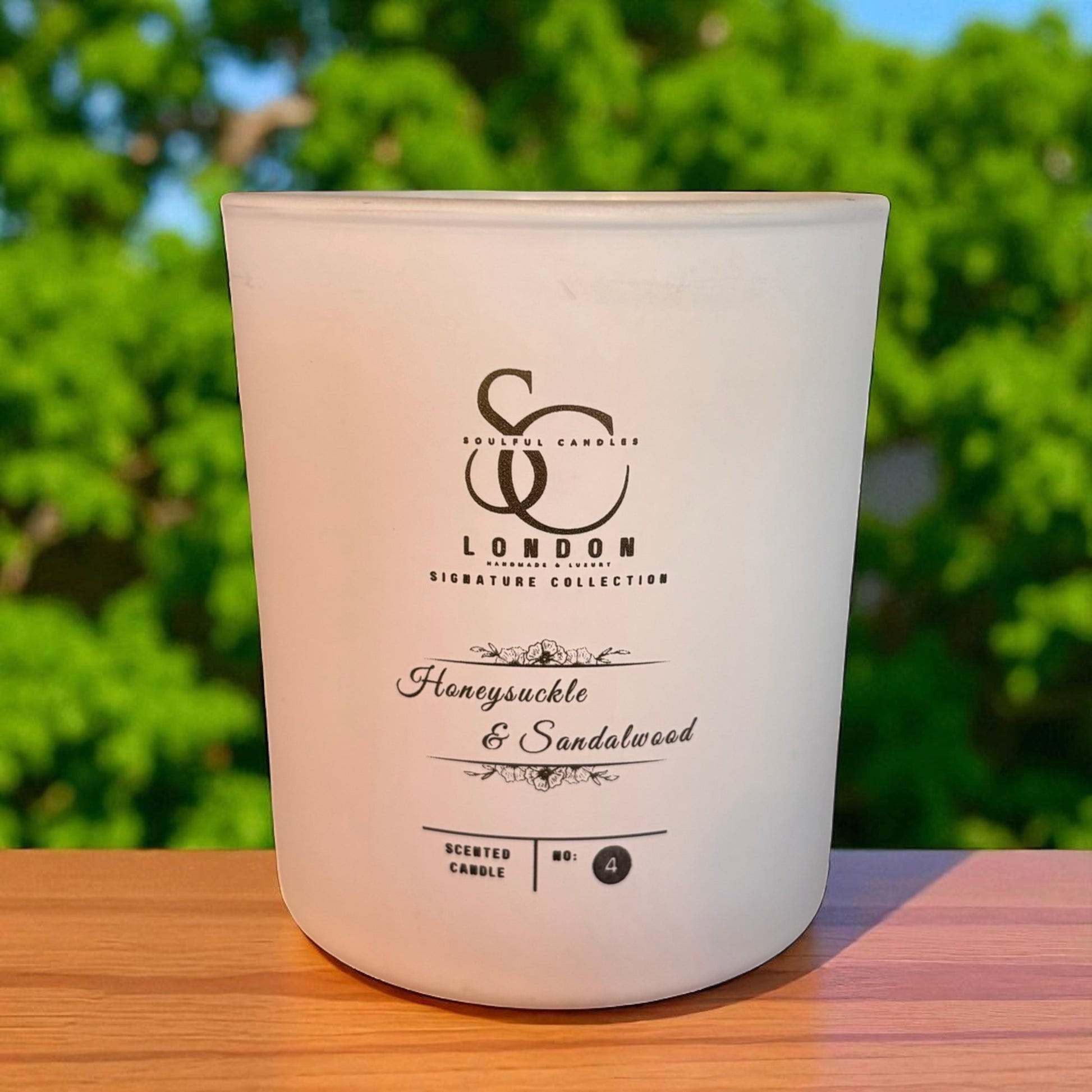 White candle with branding and scent description on a wooden surface with green foliage in the background