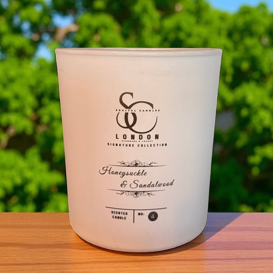 White candle with branding and scent description on a wooden surface with green foliage in the background