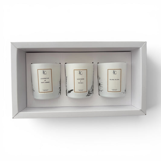 A three-scented soy candle gift set in a white rectangular box, featuring white glass jars with minimalist gold and black labels.