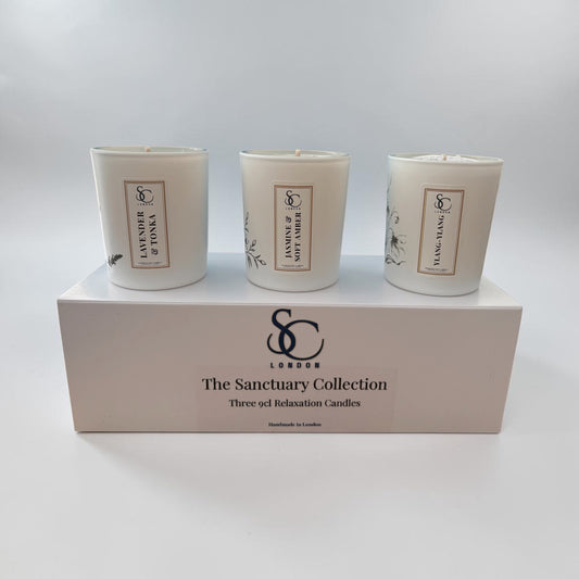 Three white scented candles in glass jars displayed on top of a white gift box labelled 'The Sanctuary Collection' with a prominent SC London logo.