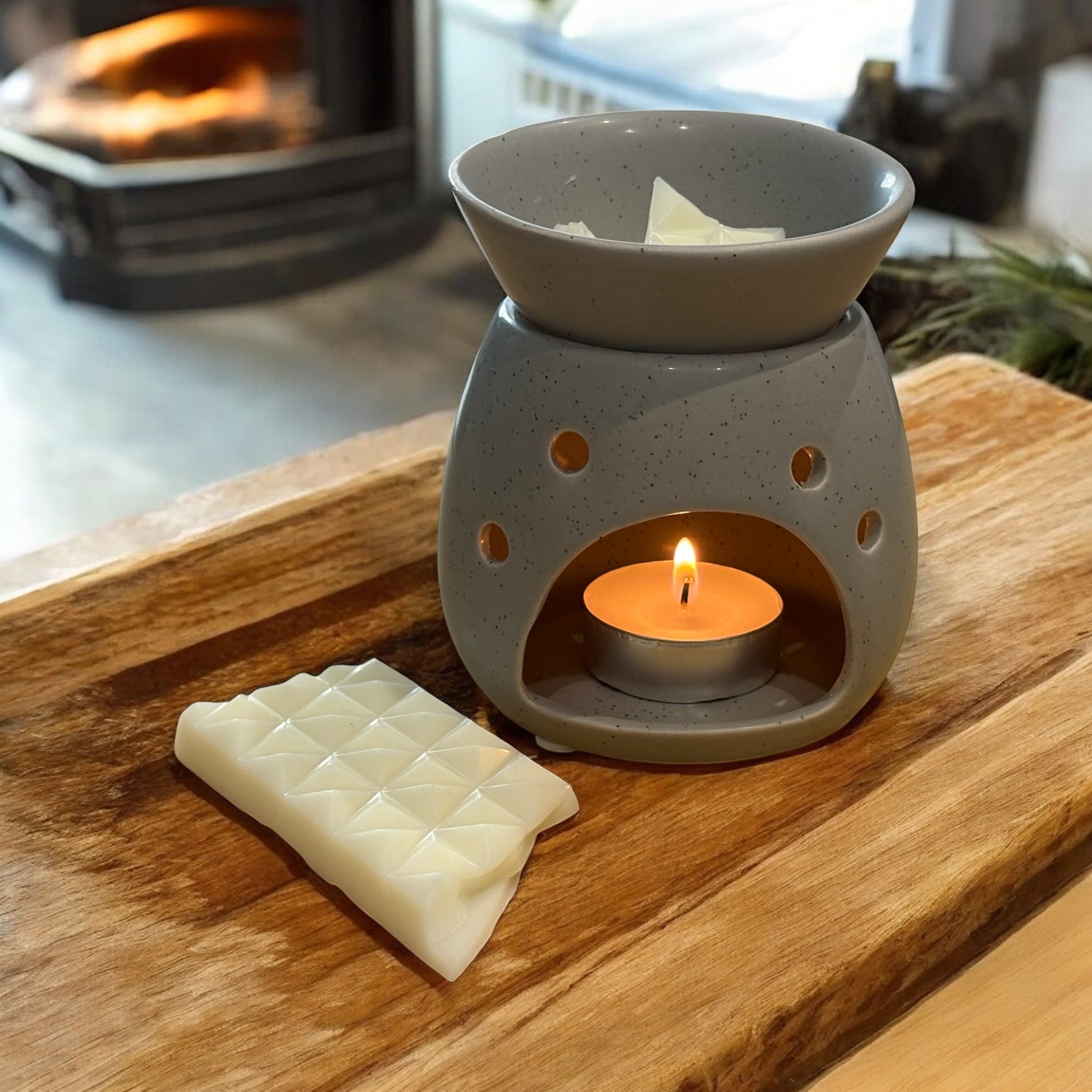 Gray ceramic wax warmer with a lit candle on a wooden surface, with a fireplace in the background.